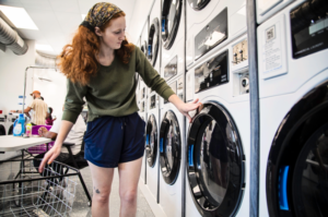 A woman with red hair, wearing a bandana and blue shorts, checks the control panel of a white front-loading washing machine in an industrial laundromat.