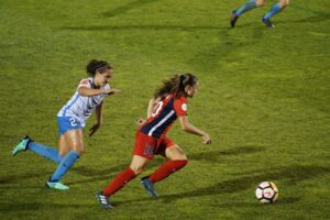 Vista aérea de una jugadora de fútbol con uniforme rojo conduciendo el balón sobre el césped, mientras es seguida de cerca por una rival con uniforme blanco y azul. Tendencias tecnológicas que están transformando la experiencia de usuario en las asociaciones recreativas.