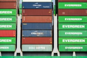 A massive stack of green, blue, and red shipping containers bearing the logos of international shipping lines such as China Shipping, Evergreen, and CMA CGM at a commercial port.