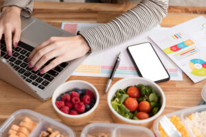Top view of a person typing on a laptop on a wooden desk, surrounded by a smartphone, printed reports, and containers of fresh, healthy food.