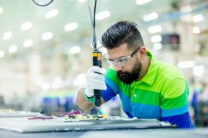 An electronics manufacturing worker carefully assembles a component onto a circuit board, using a power tool and safety equipment, inside a well-lit factory in North America.