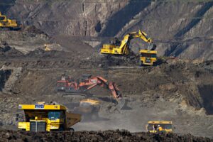 Yellow excavators and giant dump trucks operate in an open-pit mine with large earth cuts, representing the extraction and development of mineral deposits globally.