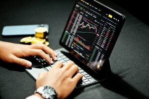 Primer plano de manos escribiendo en un teclado de tablet que muestra gráficos bursátiles y tablas de datos en tiempo real. Al fondo, un smartphone y monedas de oro físicas. Guía para inversores online: cómo elegir una estrategia para tus objetivos.