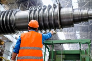 A worker wearing an orange hard hat and a reflective vest looks at a massive industrial steel rotor inside a heavy manufacturing plant equipped with high-precision technology.