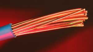 Close-up of a stripped electrical cable showing multiple strands of shiny copper emerging from blue insulation, against a warmly lit background in shades of red and dark tones.