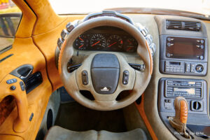 Close-up photograph taken from a low angle of a car’s interior, focusing on the leather steering wheel, the dashboard with analog gauges, and the center console with driver controls.