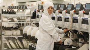 A worker wearing a sterilized protective suit operates specialized industrial machinery for electronic assembly in an advanced manufacturing plant equipped with precision components and automated instrumentation.