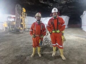 Two miners wearing overalls and hard hats pose standing inside a lit mine tunnel, with a drilling rig operating in the background.