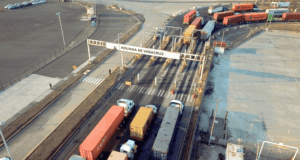 Vista aérea de carriles de peaje en la aduana de Veracruz, con camiones de carga de colores rojo y amarillo alineados para cruzar la frontera portuaria en un área industrial. Padrón suspendido con mercancía en tránsito.