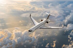 A Bombardier Global 7500 business jet, featuring a textured white livery, flies at high altitude over a sea of clouds illuminated by the evening light.