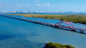 Vista aérea panorámica del Puente Vehicular Nichupté cruzando la laguna turquesa, destacando su estructura elevada y un característico arco metálico rojo rodeado de vegetación de manglar.