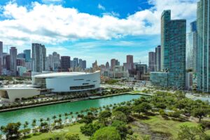Vista aérea del centro urbano de Miami con rascacielos modernos, el estadio Kaseya Center frente a la bahía de Biscayne, áreas verdes y un canal con agua turquesa bajo cielo soleado. ¿Por qué los inversionistas mexicanos están blindando su patrimonio con bienes raíces en Miami?