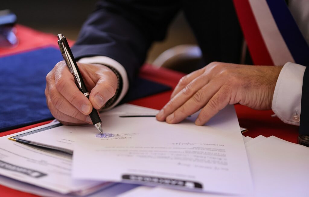 Acercamiento a las manos de un representante firmando un documento oficial con pluma negra sobre un escritorio con mantel rojo, mientras porta una banda tricolor en su traje formal.