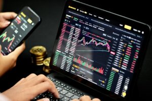 A person analyzes Japanese candlestick charts and real-time market data on a laptop and a smartphone, surrounded by gold coins on a dark desk.