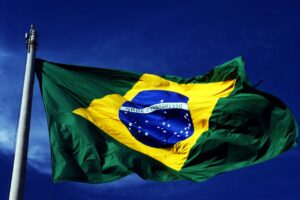 The Brazilian flag flies proudly from a metal flagpole against a deep blue sky dotted with white clouds. The words “ORDEM E PROGRESSO” are written in the center of the flag.