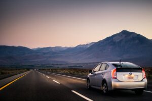 Un vehículo plateado circula por una carretera asfaltada de dos carriles, delimitada por líneas amarillas y blancas, en un entorno desértico con montañas al fondo bajo un cielo al atardecer. Checklist para preparar tu automóvil antes de realizar un viaje largo por carretera.