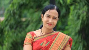 Retrato de una mujer india sonriente vestida con un sari rojo con detalles verdes y dorados, con un fondo verde natural desenfocado, representando a las emprendedoras del país.
