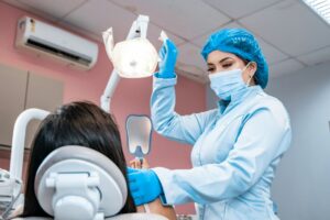 A professional dentist wearing a lab coat, face mask, and blue gloves adjusts the office lamp while treating a patient seated in the dental chair, who is holding a tooth-shaped mirror. Mexico Creates 13,469 Simplified Joint-Stock Companies (SAS) in 11 Months.