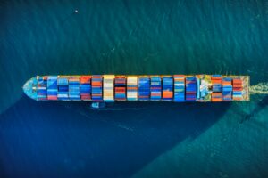 Vista aérea de un gran barco de carga repleto de contenedores multicolores navegando por el océano azul, simbolizando el transporte marítimo de mercancías y la importación de bienes de capital.