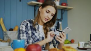 Una joven sentada a la mesa ingresa los datos de su tarjeta bancaria en su teléfono móvil mientras gestiona transacciones y compras digitales desde la comodidad de su hogar. Cómo gestionar pedidos de varios canales sin historial de crédito.
