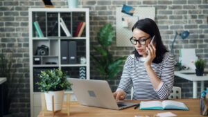 Una mujer joven en su oficina en casa, usando lentes y una camisa a rayas, mientras habla por celular y trabaja en su laptop, ilustrando la búsqueda de información crediticia en México. Tarjeta de crédito en México: claves para entender el mercado.