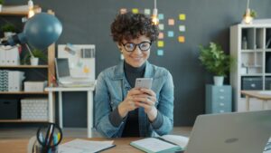 Una mujer joven sonríe mientras usa su teléfono inteligente, sentada frente a una computadora portátil en un escritorio de oficina con documentos y suministros organizados de fondo.