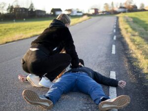 Una persona vestida de negro inclinada auxiliando a un hombre tendido en el suelo de una carretera rural de asfalto, mientras este permanece inmóvil cerca de la línea blanca.