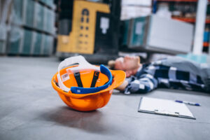 Casco naranja en primer plano en el suelo de un almacén, con un trabajador tumbado al fondo y una carretilla elevadora detrás. Los verdaderos costes de los accidentes laborales graves para empleados y empresas.