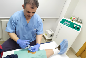 A male clinician in blue scrubs and gloves uses a syringe with a tube on a patient's draped lower leg. An advanced ozone therapy machine is visible in the background.