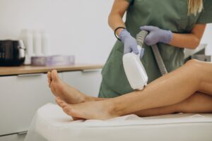 A practitioner in green scrubs and purple gloves applies a laser hair removal handpiece over transparent gel on a patient's lower leg in a modern clinical setting.