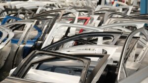 Una toma de ángulo medio muestra filas y filas de puertas de coche apiladas en un desguace bajo un cielo azul brillante. La mayoría de las puertas son blancas, pero algunas son negras, azules y rojas, lo que crea un patrón visual colorido. Productores de autopartes de Estados Unidos representan 66% del valor de un vehículo.
