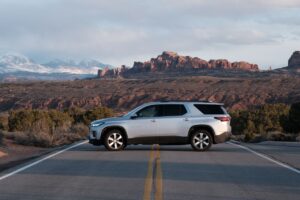 A silver SUV is parked in profile on a desert highway, with rocky mountains and snow-capped peaks in the background, symbolizing the presence and movement of the automotive industry in North America.