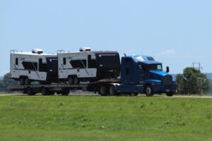 Um caminhão com cabine azul, visto de perfil em uma rodovia em um dia ensolarado, reboca um reboque de plataforma baixa com dois reboques de viagem de quinta roda brancos e pretos atrás. As 6 propostas da PACCAR sobre a revisão do T-MEC.