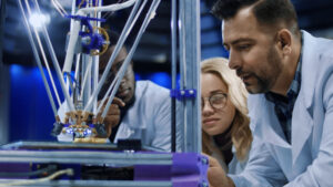 Tres ingenieros con batas laboratorio observan atentos el proceso de impresión 3D de alta precisión con tecnología avanzada, analizando detalles técnicos del prototipo industrial en desarrollo tecnológico. Ventaja competitiva industrial: la visión de Flex.